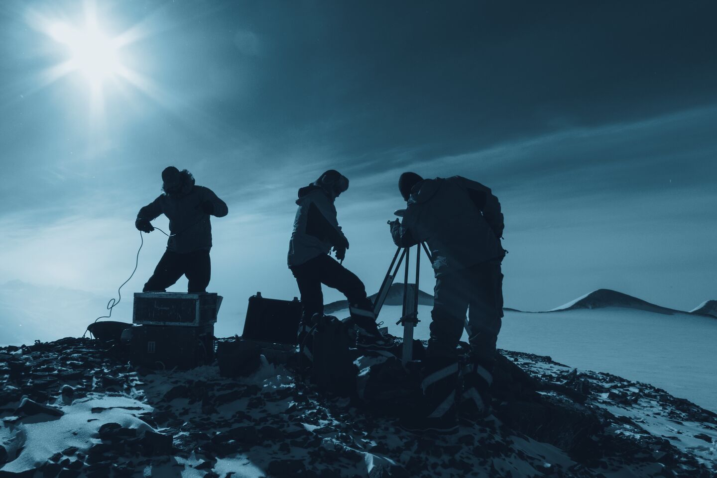 Three people working with survey equipment on a rocky summit above an ice sheet, backlit by a low sun.
